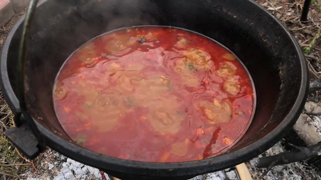 Making red soup in cast iron container with little bonfire under it. Making food and eating while camping in nature. Bubbles in boiling soup. Survival mode.