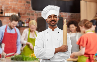cooking class, profession and people concept - happy male indian chef or baker in toque with rolling-pin over group of students background