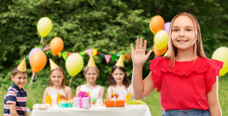 childhood, greeting and people concept - beautiful smiling girl in red shirt and skirt waving hand at birthday party over friends in summer park background