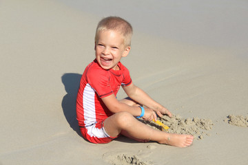 A little blond boy in clothes with UV filter is playing with sand on the beach by the sea, holiday with children, protecting kids from the sun concept