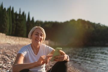 Mature woman sitting after jogging on beach. Senior lady using smartphone and earphones at sunset. Evening running