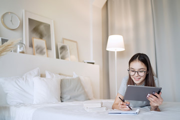 Cute teenager girl doing homework lying on bed at home. Young pretty girl wearing glasses writing...