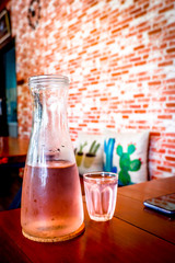 Cool water in tall jar with small glass on the wooden table and brick wall in cafe