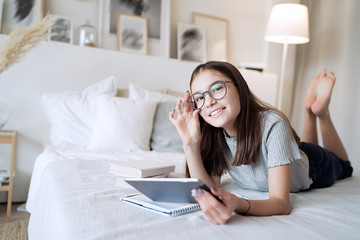 Cute teenager girl using tablet lying on bed at home. Young pretty girl wearing glasses studying online, distance learning, self education