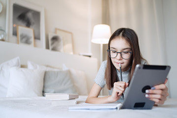 Cute teenager girl doing homework lying on bed at home. Young pretty girl wearing glasses writing down in notebook, studying online with tablet, distance learning, self education     