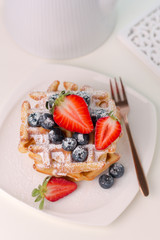 Stack of delicious waffles with blueberries and sliced strawberries .close-up on white table . with white teapot from the back