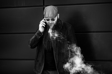 Vape bearded man. An adult man with a very long beard in a cap and sunglasses smokes an electronic...