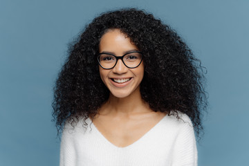 Indoor shot of lovely curly woman with pleasant smile, wears spectacles and white casual jumper, isolated over blue background, expresses good emotions. Feminine girl enjoys nice day with friends