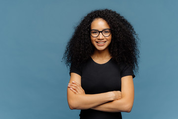 Waist up shot of smiling Afro American woman has arms folded, wears spectacles and casual black t...