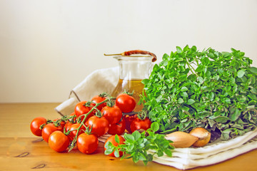 Cherry tomatoes on a sprig next to the greens of fresh basil, shallot onions, chili pepper, olive oil in a transparent decanter on a white napkin on a wooden table. Copy space.