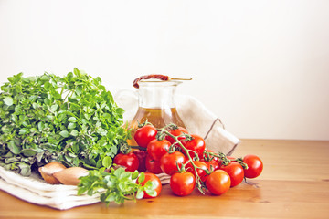 Cherry tomatoes on a sprig next to the greens of fresh basil, shallot onions, chili pepper, olive oil in a transparent decanter on a white napkin on a wooden table. Copy space.