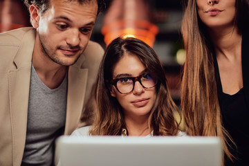 Charming buisnesswoman with brown hair and dressed elegant sitting at restaurant and looking at laptop. Next to her standing two colleagues and looking at laptop, too.