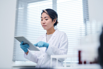 Serious busy young Asian female laboratory specialist in sterile gloves and white coat standing in...