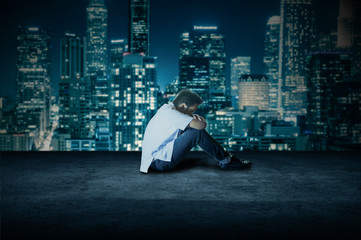 Wide shot of a young man sitting and crying on a floor of the roof top of a corporate building. Side view shot of a student very depressed. Depressed teenage boy sitting on ground thinking at night.