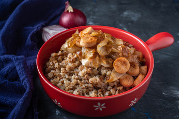 buckwheat porridge with sausages and fried onions in a red pan on a dark background, copy space, Close-up