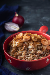 buckwheat porridge with sausages and fried onions in a red pan on a dark background, copy space, Close-up