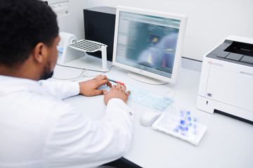 Over shoulder view of concentrated black experimental scientist in white coat sitting at table and typing on computer while recording data of research