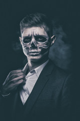 Fototapeta premium Stylish and beautiful, emotional young man with skeleton makeup in a strict suit against the background of smoke and dark background to Halloween