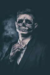 Stylish and beautiful, emotional young man with skeleton makeup in a strict suit against the background of smoke and dark background to Halloween