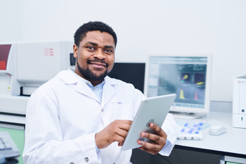 Content confident young black medical expert with beard and mustache sitting at desk with modern computer and using digital tablet in laboratory