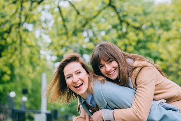 Fototapeta premium Two women friends laughing and hugging outdoors. Adult playfull good looking women sisters in trendy coat piggybacking her pretty girlfriend in spring park outdoor. Real emotion, true friendship