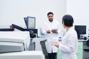 Serious young black doctor in lab coat standing at desk and using tablet while talking to colleague...