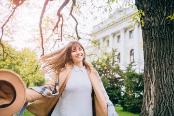 Outdoors lifestyle portrait of brunette hair woman with loose hair in the wind in beige hat and trendy coat walking at city park . Female whirling. Cold spring weather season concept.