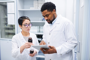 Serious thoughtful multi-ethnic pharmacists in lab coats standing in drugstore and discussing new...