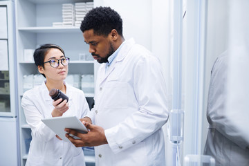Fototapeta premium Young Asian female medical assistant in glasses showing pill bottle to black doctor and asking him about pills in storage room