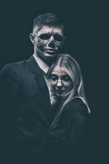 Stylish and beautiful, emotional young man with skeleton makeup in a strict suit against the background of smoke and dark background to Halloween hugs slender, beautiful blonde with glasses and black 