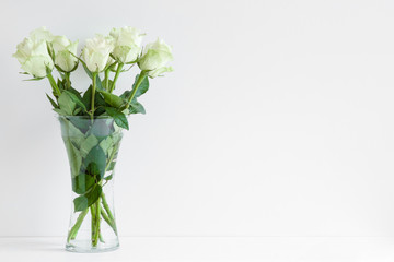 White, light green roses in glass vase on table at light gray wall. Mockup for positive idea. Empty...