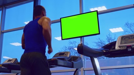Strong athletic man running on a treadmill at a gym and watching a green screen