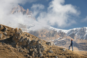 A traveler is a man with a beard and a hat standing on mountain range at the foot of epic rocks partly hidden by clouds in the mountains an adventure travel vacation in Norway or the Caucasus