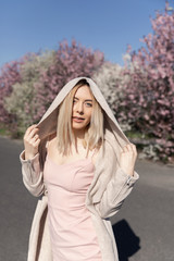 Young beautiful blonde girl stands in the parking lot. Spring flowering trees.