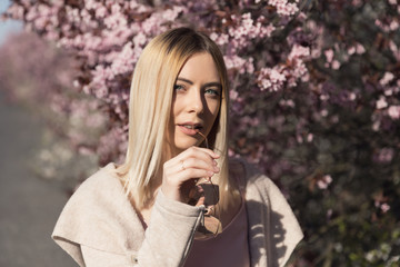 Young beautiful blonde girl stands in the parking lot. Spring flowering trees.