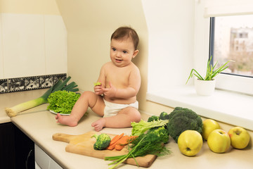 The baby in the diaper is sitting on the kitchen table. A child plays and has fun with fresh organic vegetables and fruits.