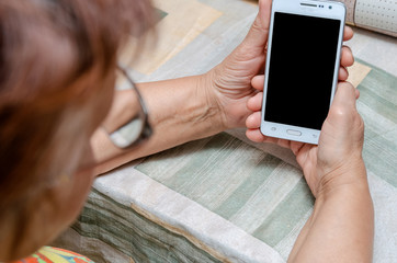 Elderly woman with glasses holding a white smartphone with black screen while sitting at the table in the kitchen, photo from the shoulder