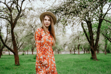 Young beautiful blonde woman in blooming garden. Spring trees in bloom. Orange dress and straw hat.