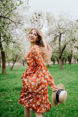 Young beautiful blonde woman in blooming garden. Spring trees in bloom. Orange dress and straw hat.