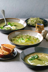 Variety of green vegetable soup asparagus, broccoli and pea, decorated by greens, vegetables, quail eggs, cream, olive oil, in ceramic bowls over grey spotted table with ingredients above.
