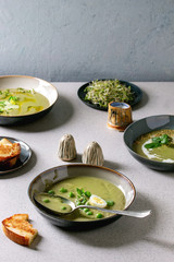 Variety of green vegetable soup asparagus, broccoli and pea, decorated by greens, vegetables, quail eggs, cream, olive oil, in ceramic bowls over grey spotted table with ingredients above.