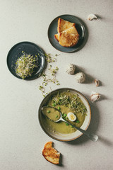 Started eaten green vegetable soup asparagus, broccoli or pea, decorated by green pea, sprouts and quail eggs in ceramic bowls with bread, salt and pepper over grey spotted background. Flat lay, space