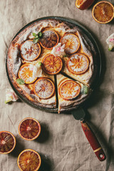 Cutted Homemade Cheesecake with sicilian blood oranges, decorated by edible flowers, mint leaves, sugar powder served in plate with cutted oranges above over grey linen table cloth. Flat lay, space