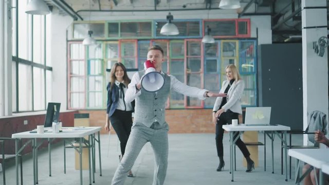 A young businessman goes to the office and starts dancing, holding a megaphone. Colleagues join, dance with him and blow up flappers with confetti. Creative coworking. Office holiday. Corporate party