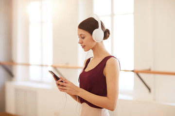  Content inspired pretty girl in wired headphones standing in empty ballet classroom and choosing track on smartphone for training