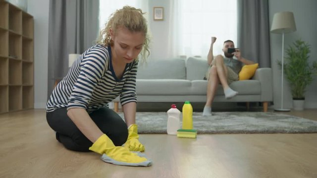 Tired wife cleans parquet floor and her husband play on smartphone on sofa