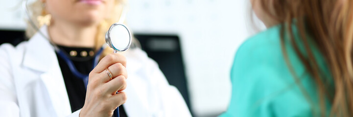 Blond mature gp hold stethoscope head in hand while diagnosing little girl