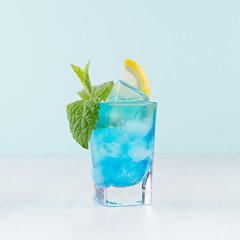 Tropical beach alcohol blue drink  - curacao liquor, lemon slice, ice, mint in misted shot glass on green background, white wood board.
