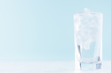 Cold water with ice in misted drinking glass on soft light blue background, white wood board.
