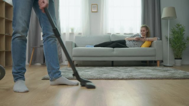 Man vacuuming parquet floor, and his girlfriend using smartphone on couch